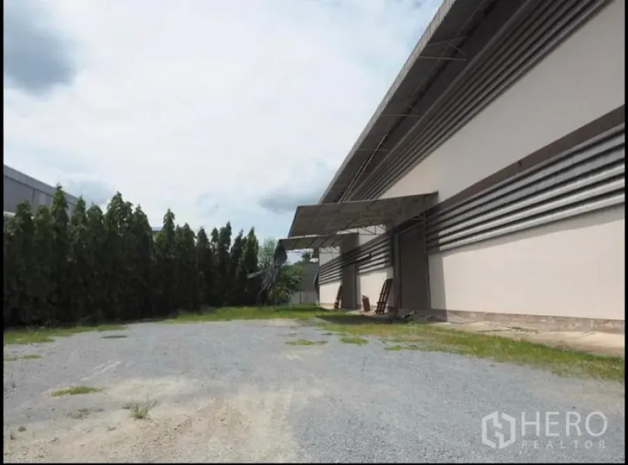 Warehouse for rent in Nakhon Pathom - Gravel yard beside a long warehouse facade with covered loading bay and tall trees along the boundary.