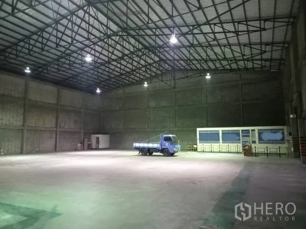 货栈 出租在Mueang Pathum Thani, Pathum Thani - 仓库内部空间开阔，钢桁架屋顶、挑高层高、照明明亮，混凝土地面上停有卡车。