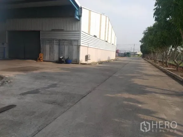 Warehouse for rent in Pathum Thani - Side view of the warehouse loading area with roller shutters and a tree-lined drive.