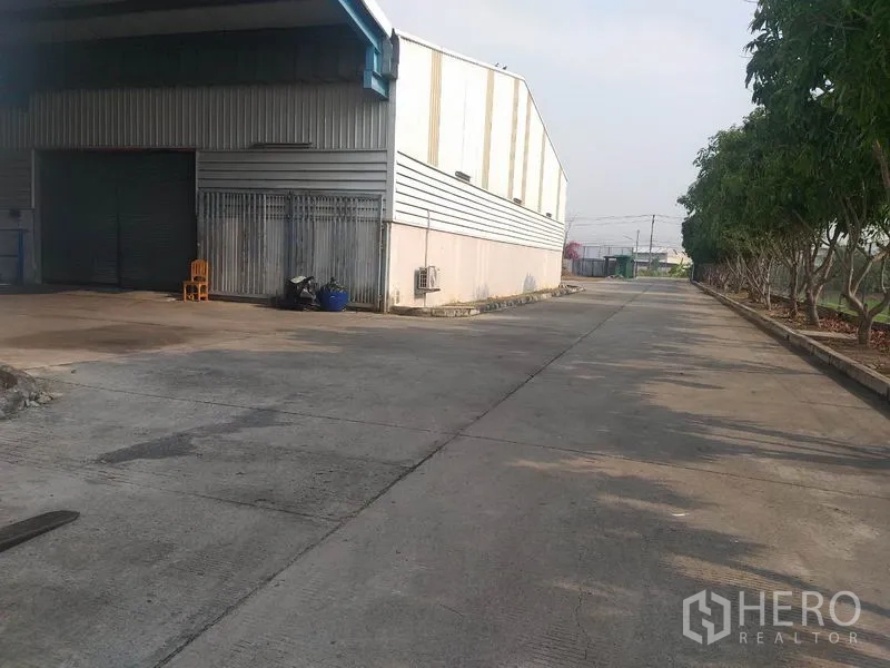 Warehouse for rent in Pathum Thani - Side view of the warehouse loading area with roller shutters and a tree-lined drive.