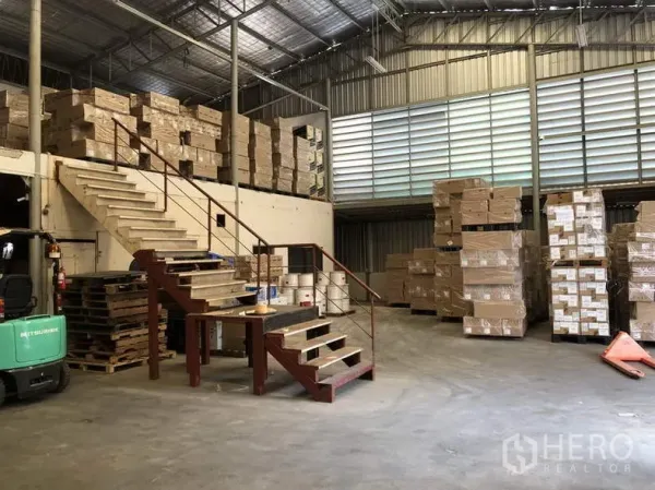 Warehouse for rent in Suan Luang, Bangkok - Spacious warehouse with louvered ventilation, mezzanine stairs, and stacked cartons on pallets.