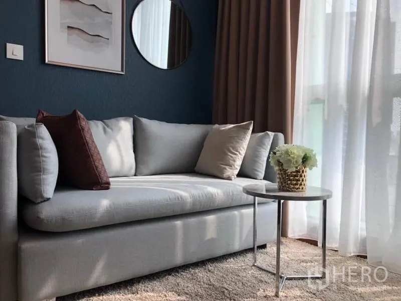 Condominium for rent or sale in Ratchathewi, Bangkok - Cozy gray sofa by large window with sheer curtains, round mirror, and side table on a soft rug.