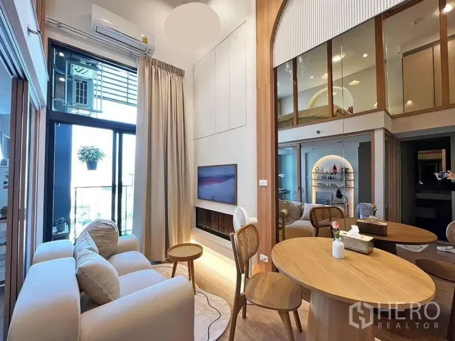 คอนโด ให้เช่าในHuai Khwang, Bangkok - ห้องนั่งเล่นเพดานสูงสองชั้น ประตูระเบียง ทีวีติดผนัง โซฟาโทนอุ่น และโต๊ะกลม