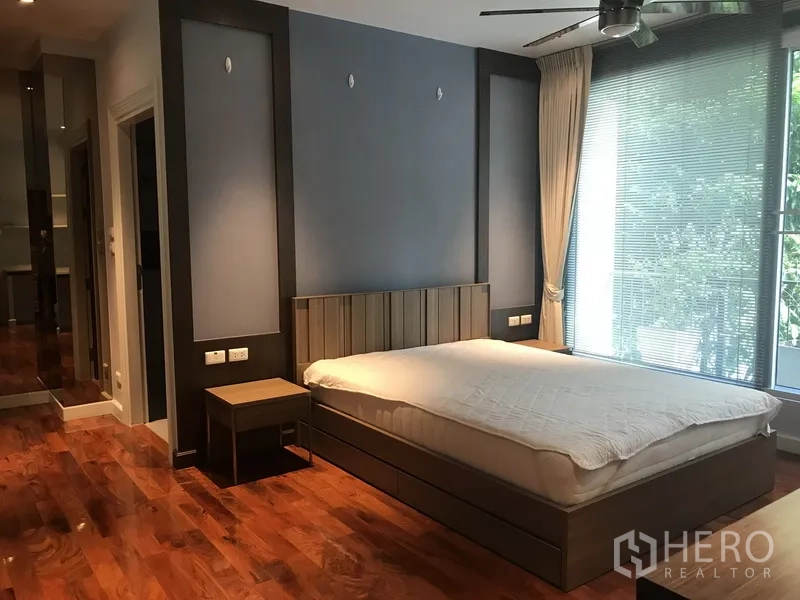 Apartment for rent in Pathum Wan, Bangkok - Large bedroom with blue feature wall, ceiling fan and full-height window with blinds.
