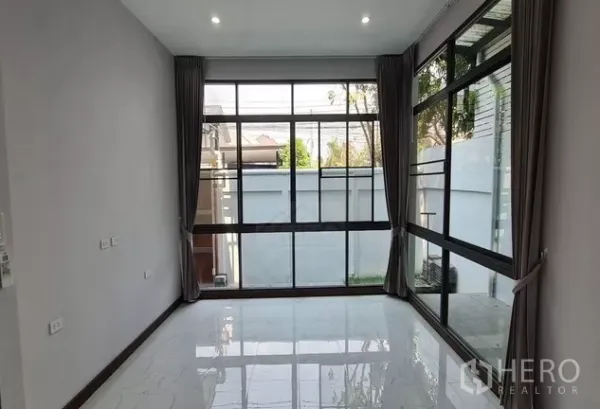 Home Office for rent in Watthana, Bangkok - Ground‑floor room with floor‑to‑ceiling corner windows, gray curtains and a view to a small garden and boundary wall.
