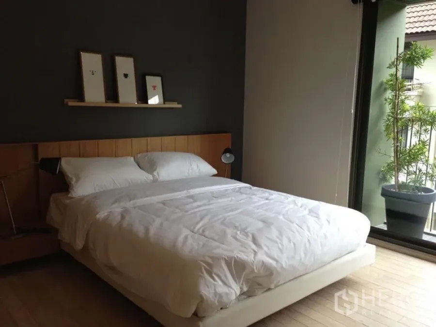 公寓 出租在Watthana, Bangkok - 明亮卧室，双人床与深色背景墙，置物架装饰画，滑门通往带绿植的阳台。