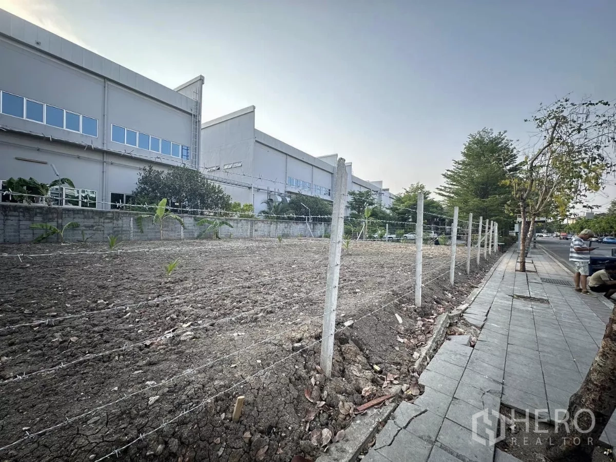 Land for sale in Saphan Sung, Bangkok - Corner of a vacant plot with barbed-wire fence along a paved sidewalk and road.
