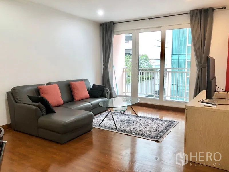 Condominium for rent or sale in Watthana, Bangkok - Living room with L-shaped leather sofa, rug, and floor-to-ceiling windows opening to a small balcony.