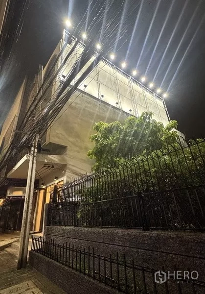 อาคารพาณิชย์ ให้เช่า - มุมมองจากทางเท้า เห็นด้านหน้าอาคารพร้อมไฟส่องป้ายดาดฟ้า รั้วเหล็ก และทางเดิน เขตพระโขนง