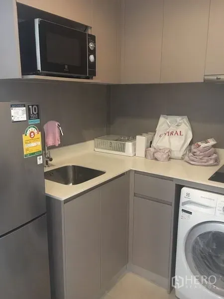 Condominium for sale - Compact L-shaped kitchen with grey cabinets, sink, microwave, refrigerator, and in-unit washing machine.