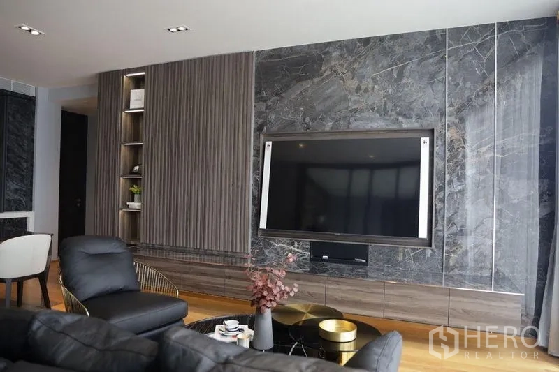 Condominium for rent in Khlong Toei, Bangkok - Contemporary living room with a large TV mounted on a marble wall, built-in shelves, leather sofa and wood floors.
