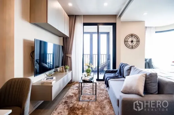 Condominium for rent in Watthana, Bangkok - Bright living room with wall-mounted TV, grey sofa and sliding glass doors to a small balcony.