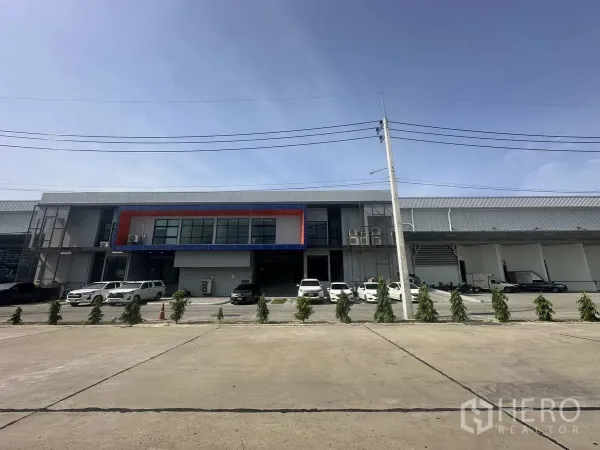 Warehouse for rent in Khlong Luang, Pathum Thani - Head-on view of the warehouse exterior with central office block, parked vehicles, and landscaped shrubs.