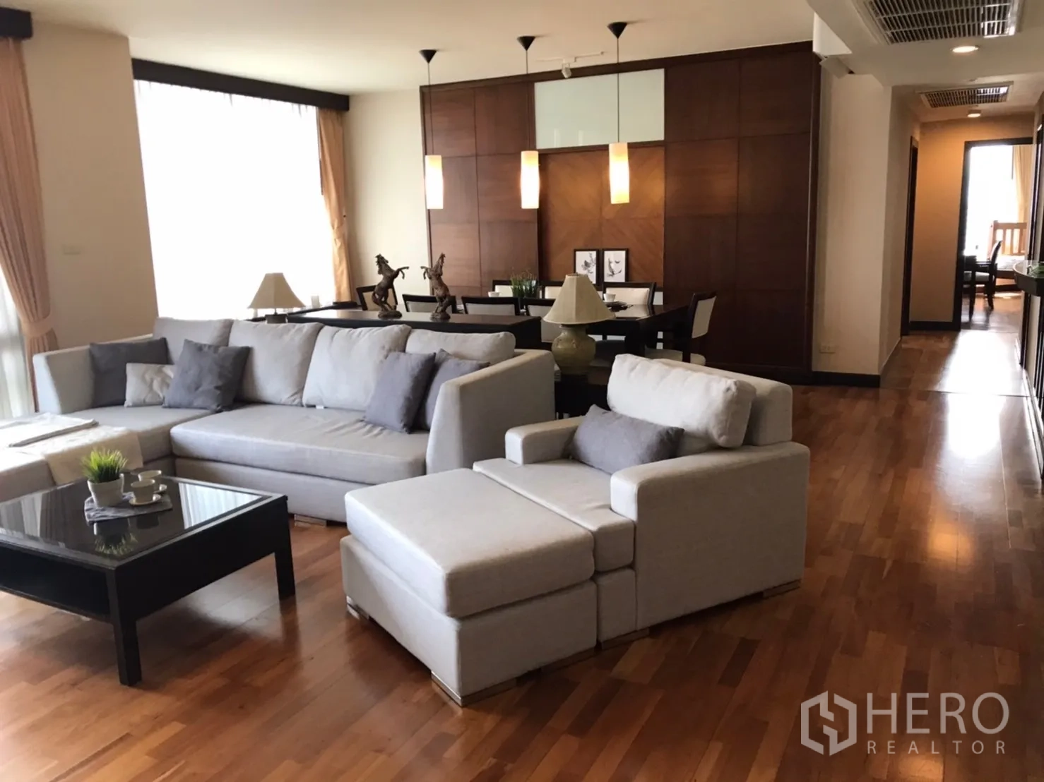 Condominium for rent or sale in Pathum Wan, Bangkok - Bright living room with L‑shaped grey sofa, black coffee table, wood floors, and dining area with pendant lights.