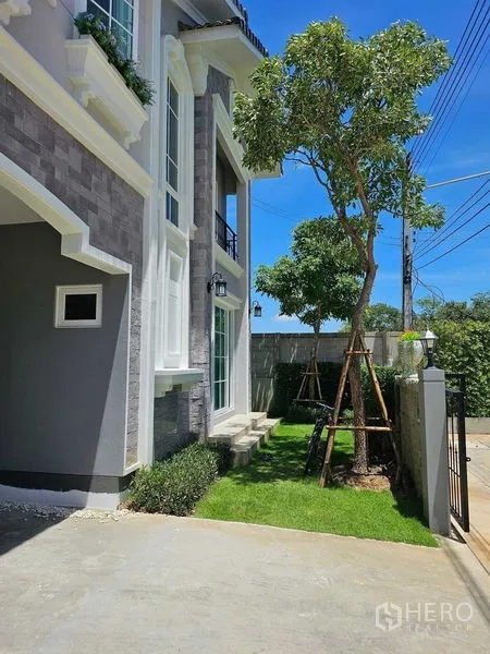 Detached House for rent in Thanyaburi, Pathum Thani - Front driveway with private gate, tree and trimmed hedges beside the home