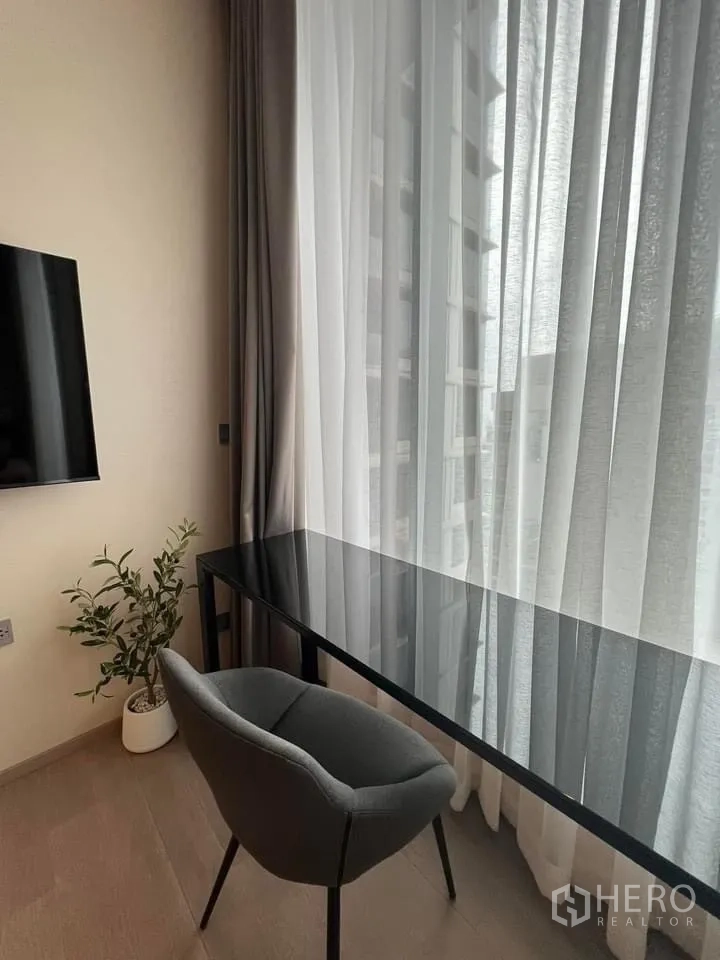 คอนโด ให้เช่า - มุมทำงานที่บ้าน โต๊ะกระจก เก้าอี้สีเทา พร้อมกระถางต้นไม้ ข้างหน้าต่างผ้าม่านสูง