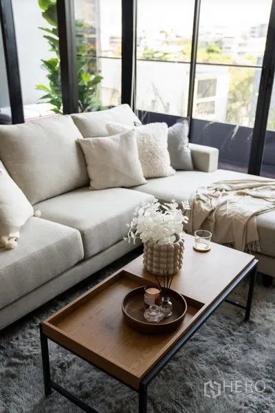 Condominium for rent or sale in Phra Khanong, Bangkok - Close-up of plush sofa and wooden coffee table with decorative tray on a soft rug beside balcony windows.