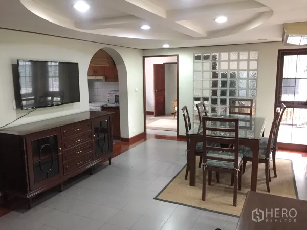 Detached House for rent in Khlong Toei, Bangkok - Dining area with glass-top table, TV console and feature ceiling beside a glass-block wall.