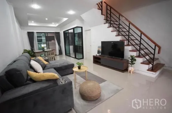 Townhouse for rent in Bang Kapi, Bangkok - Open-plan living room with grey sectional sofa, TV console and wooden staircase beside a glass partition.