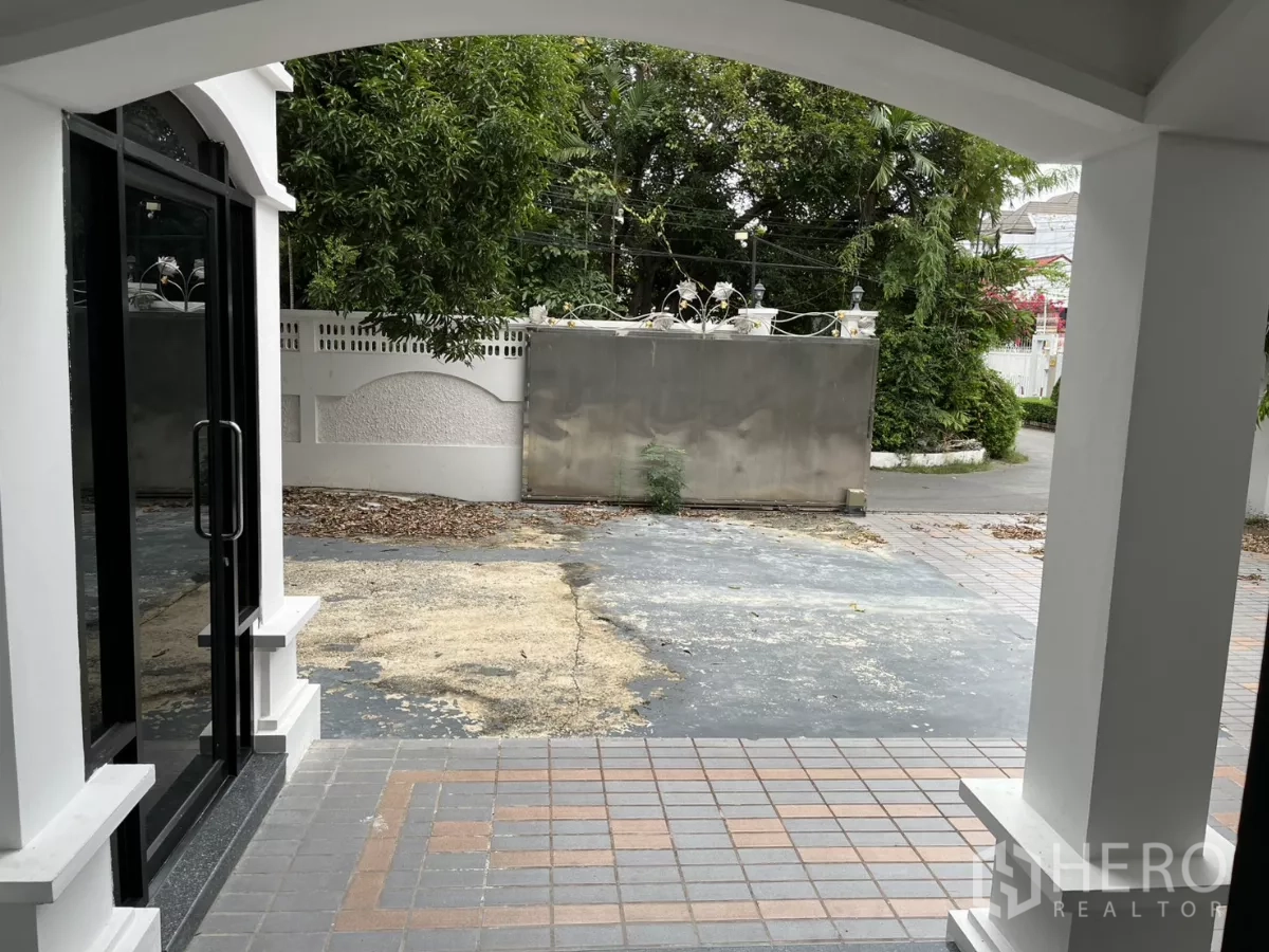 บ้าน ให้เช่าในWatthana, Bangkok - มุมมองจากเฉลียงสู่ลานหน้าบ้านปูพื้นกระเบื้อง พร้อมประตูรั้วและต้นไม้ใหญ่.