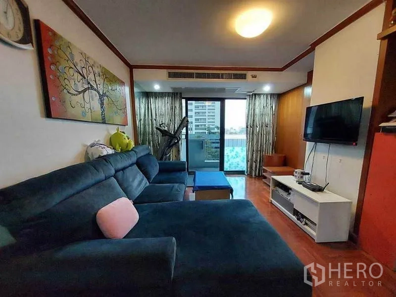 Condominium for rent or sale in Watthana, Bangkok - Living room with dark sectional sofa, wall-mounted TV and sliding doors to a balcony with city view.