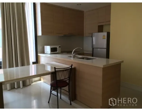 Condominium for rent in Khlong Toei, Bangkok - Modern light-wood kitchen with island sink, microwave, refrigerator and dining nook by a tall window.