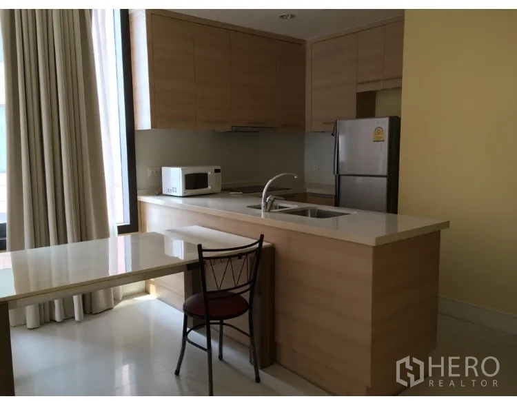 Condominium for rent in Khlong Toei, Bangkok - Modern light-wood kitchen with island sink, microwave, refrigerator and dining nook by a tall window.