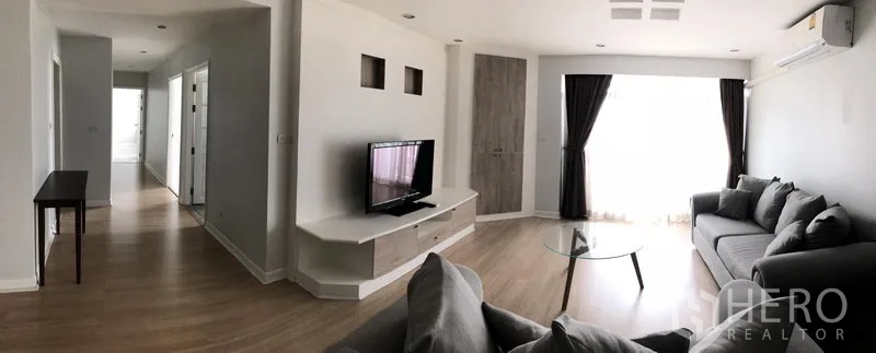 公寓 出租在Watthana, Bangkok - Royal Castle公寓明亮客厅，灰色沙发、电视柜、木地板与大窗帘落地窗。