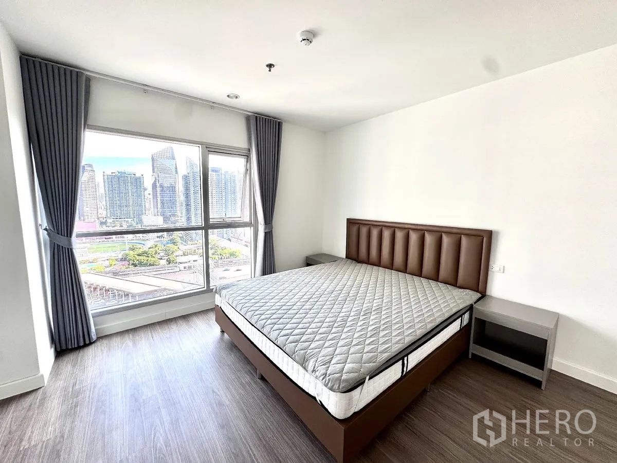 Condominium for rent in Huai Khwang, Bangkok - Second bedroom with wide window, bed, side tables, curtains, and a skyline outlook.