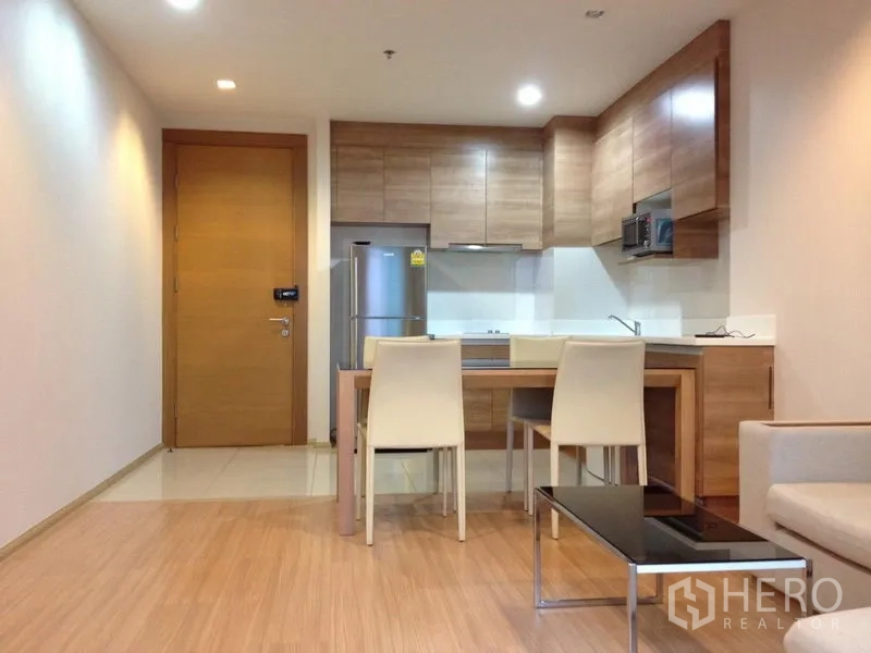 Condominium for rent in Huai Khwang, Bangkok - Open-plan living room facing the kitchen, with dining table, chairs, and a sleek coffee table on warm wood floors.