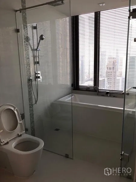 Condominium for rent in Sathon, Bangkok - Contemporary bathroom with glass shower, bathtub beside a window with blinds and city views.