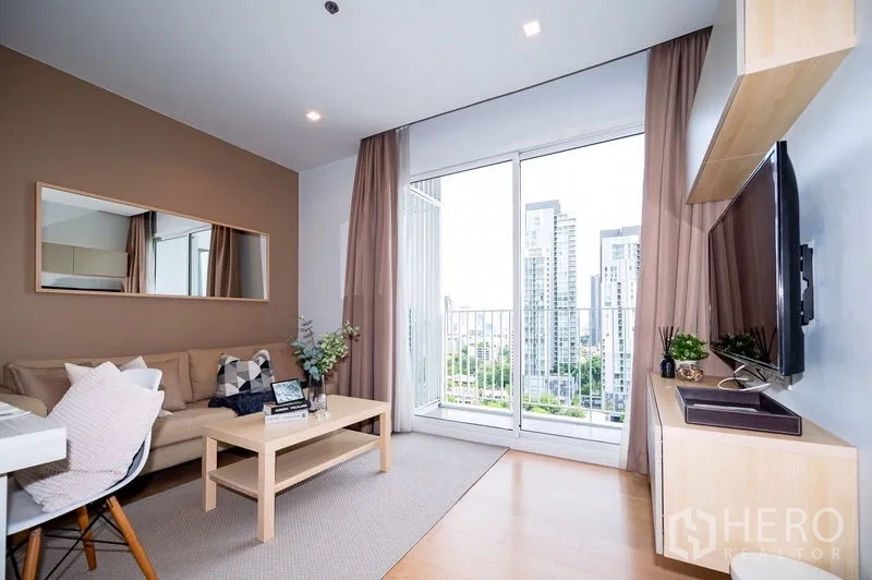 Condominium for rent or sale in Watthana, Bangkok - Cozy living room with sofa, wall mirror, and balcony access framing a high-rise city outlook.