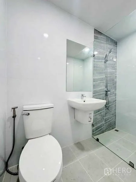 Townhouse for rent in Min Buri, Bangkok - Modern bathroom with glass shower, white sink and toilet, and gray tiled accent wall.