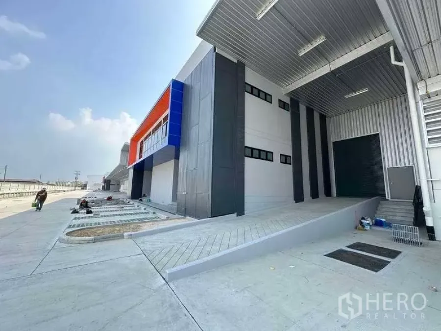 Warehouse for rent in Bang Phli, Samut Prakan - Modern warehouse exterior with covered loading ramp, orange-blue facade and wide driveway.