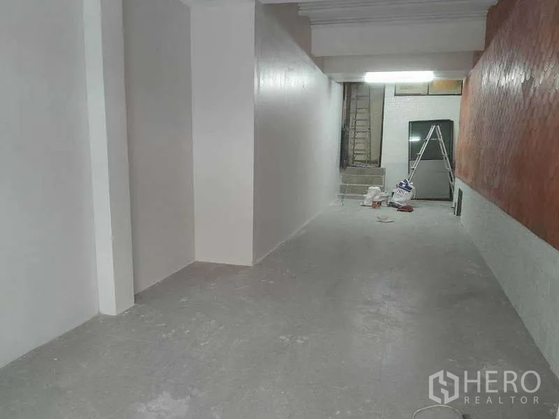 零售空间 出租在Watthana, Bangkok - 狭长的空置商铺内部，白色墙面、灰色地面与顶灯，后方有出入口和装修工具。