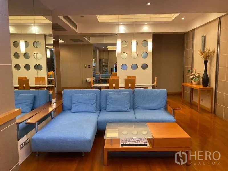 Condominium for rent in Pathum Wan, Bangkok - Open‑plan lounge with a blue sectional sofa, mirrored wall and warm lighting.