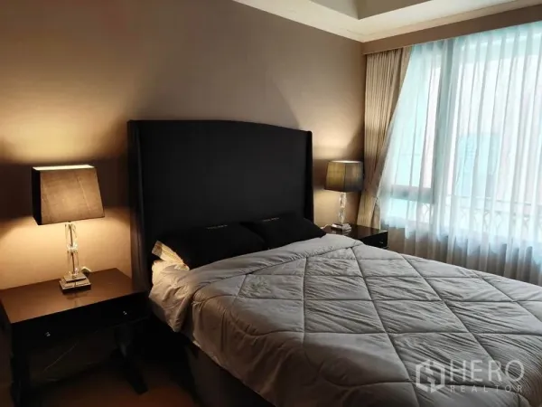Condominium for rent or sale in Pathum Wan, Bangkok - Cozy bedroom with dark upholstered headboard, bedside lamps, and a large window with sheer drapes.