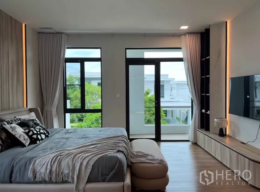 Detached House for rent in Samut Prakan - Spacious master bedroom with balcony doors, floor-to-ceiling windows, TV wall unit and wood floors.