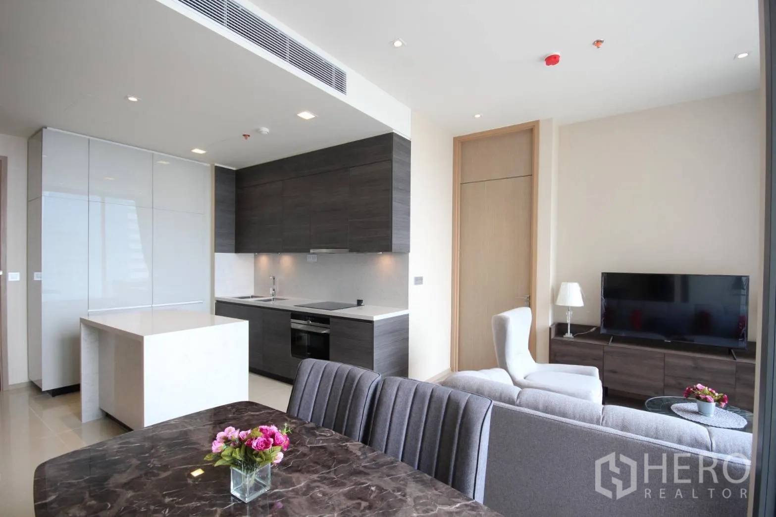 Condominium for rent in Watthana, Bangkok - Open-plan living room with gray sofa, TV console, and modern kitchen with island at The Esse Asoke.