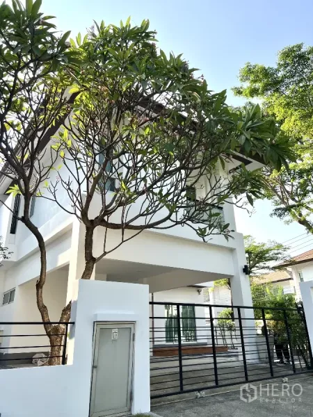 บ้าน ให้เช่าหรือขายในSaphan Sung, Bangkok - มุมหน้าบ้านแสดงโรงจอดรถ ประตูรั้วเหล็ก และต้นลีลาวดีใหญ่