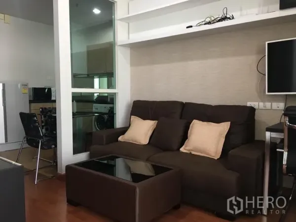 公寓 出租在Pathum Wan, Bangkok - 温馨客厅，棕色沙发、玻璃茶几、壁挂置物架，窗旁连向厨房。