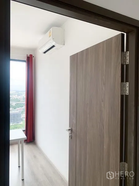 Condominium for sale in Bang Na, Bangkok - Small room with wall-mounted air conditioner, window view, and red curtains.