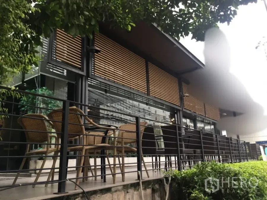 零售空间 出租在Khlong Toei, Bangkok - 玻璃立面与木百叶的现代店铺外观，设有绿树环绕的露台座位区。