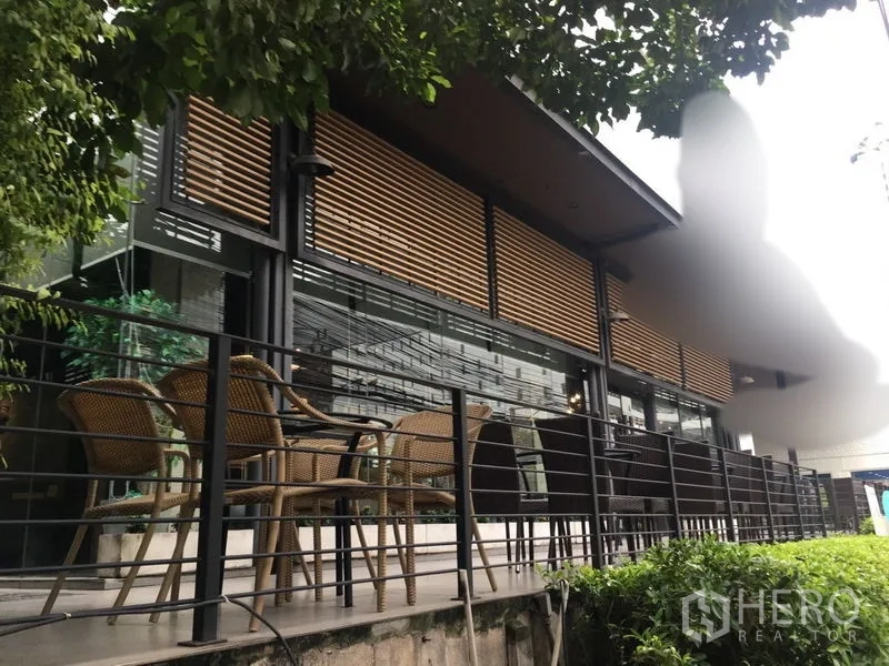 零售空间 出租在Khlong Toei, Bangkok - 玻璃立面与木百叶的现代店铺外观，设有绿树环绕的露台座位区。