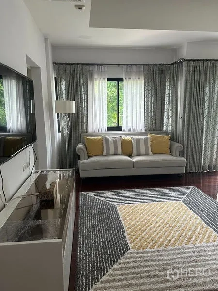 Detached House for sale in Saphan Sung, Bangkok - Light-filled family lounge with sofa, TV wall, patterned rug and sheer curtains.