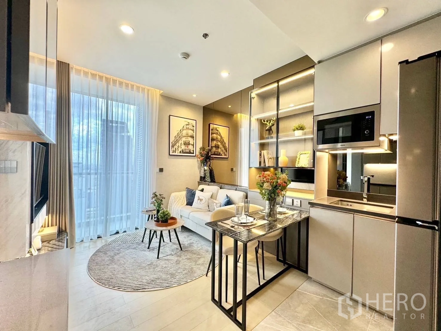คอนโด for rent in Ratchathewi, Bangkok - ห้องนั่งเล่นสว่างด้วยหน้าต่างสูงจรดเพดาน พรมทรงกลม และชั้นวางบิวท์อินข้างครัวดีไซน์เรียบหรู