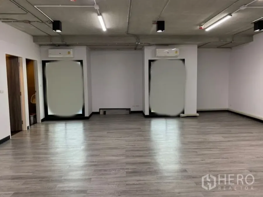 Office Space for rent - Open-plan office with grey wood-look flooring, exposed concrete ceiling, LED strip lighting, and two large windows with wall air conditioners.