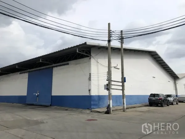 Warehouse for rent in Samut Prakan - Corner view of blue-and-white warehouse with sliding door, parking bays and overhead power lines.