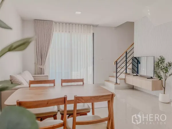 Townhouse for rent in Bang Phli, Samut Prakan - Dining table facing sheer-draped sliding doors with TV console and stairs in a light-filled living room.