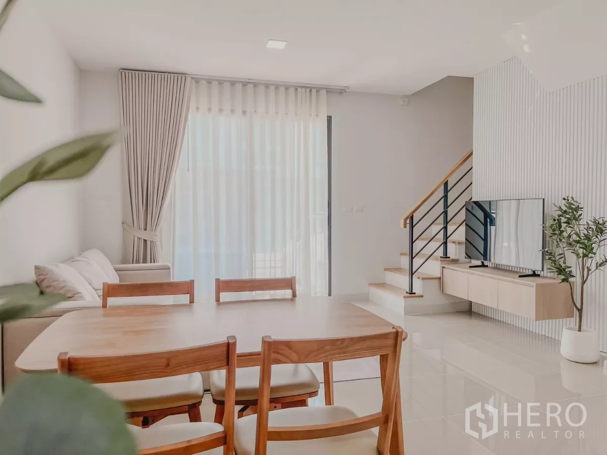 Townhouse for rent in Bang Phli, Samut Prakan - Dining table facing sheer-draped sliding doors with TV console and stairs in a light-filled living room.